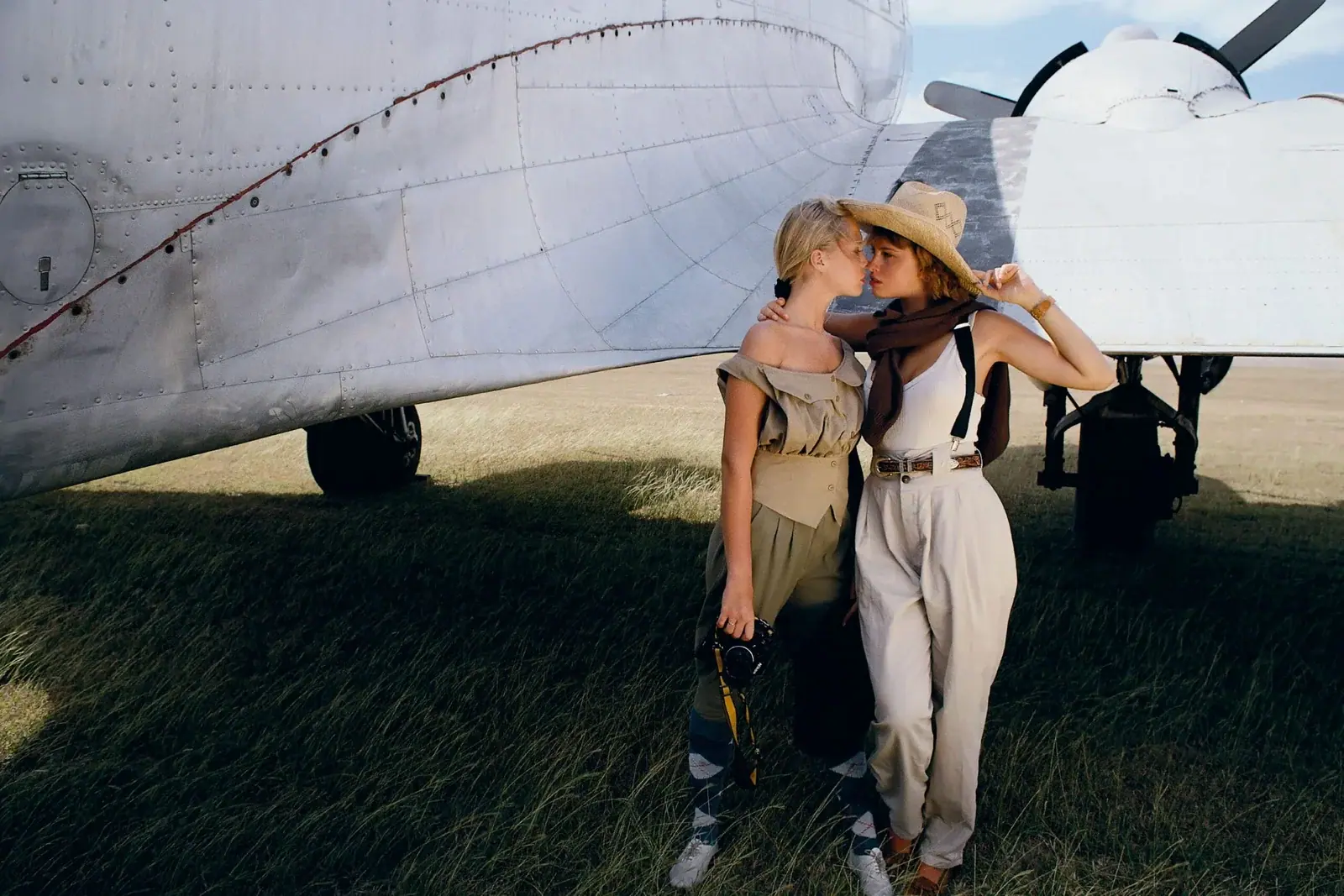 Deux femmes élégamment habillées dans un style vintage (tenues de voyage des années 40-50) se tenant près d'un avion ancien sur un terrain herbeux, sur le point de s'embrasser.